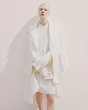 Model standing against a plain light wall wearing an off-white double-breasted blazer and layered knitwear
