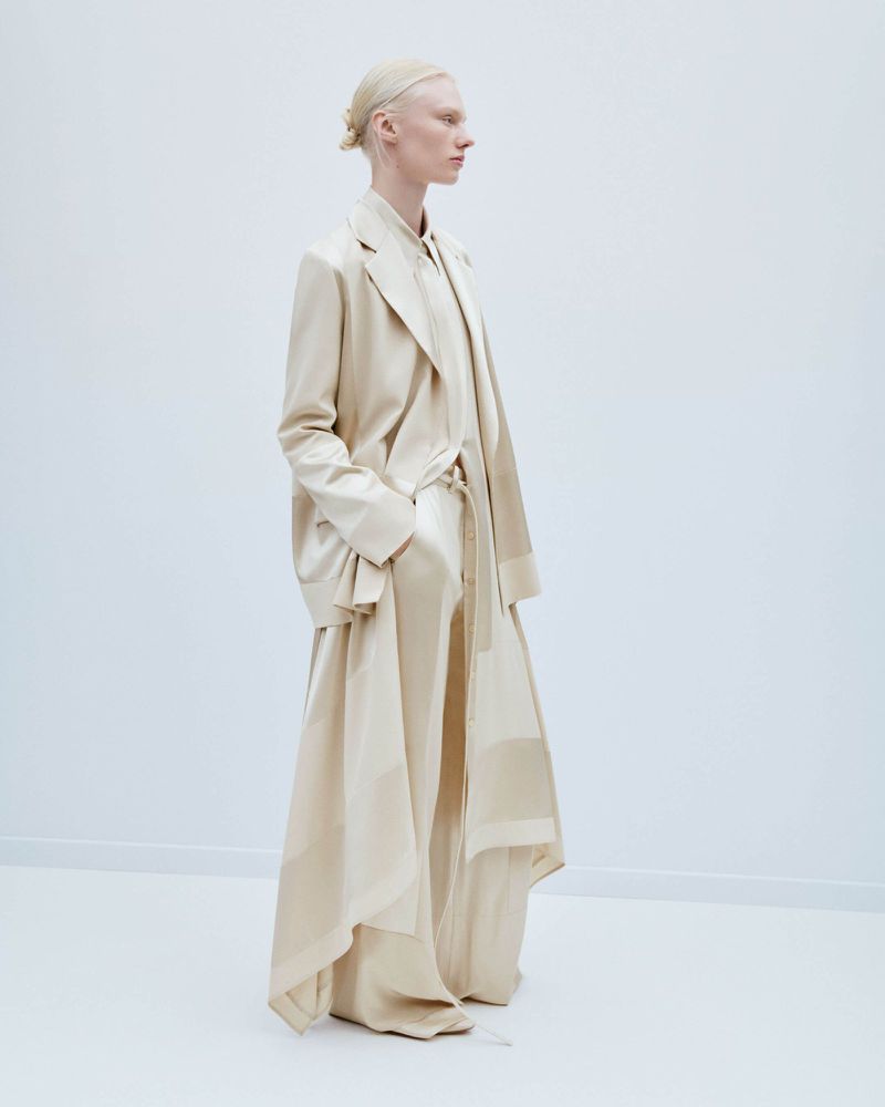 Fashion shot of a slender adult female with blonde hair pulled back, wearing a cream-colored coat and long dress against a plain white backdrop.