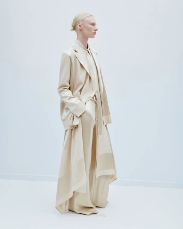 Fashion shot of a slender adult female with blonde hair pulled back, wearing a cream-colored coat and long dress against a plain white backdrop.