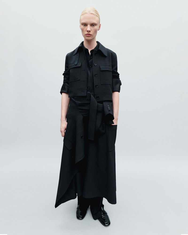 Full-length studio shot of a fair-skinned woman in a stylish all-black layered outfit with a jacket and a long skirt against a white background.