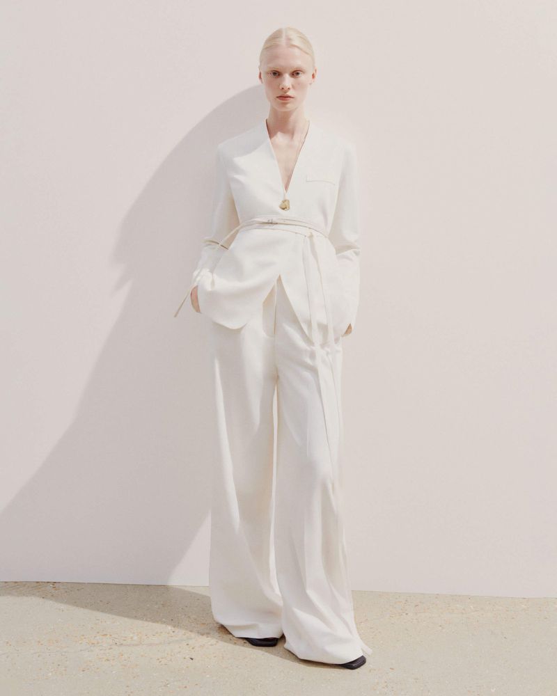 A blonde female model poses in a white blazer and wide-leg pants against a light background, casting a soft shadow on the wall.