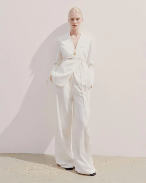 A blonde female model poses in a white blazer and wide-leg pants against a light background, casting a soft shadow on the wall.