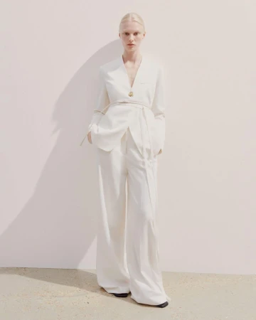 A blonde female model poses in a white blazer and wide-leg pants against a light background, casting a soft shadow on the wall.
