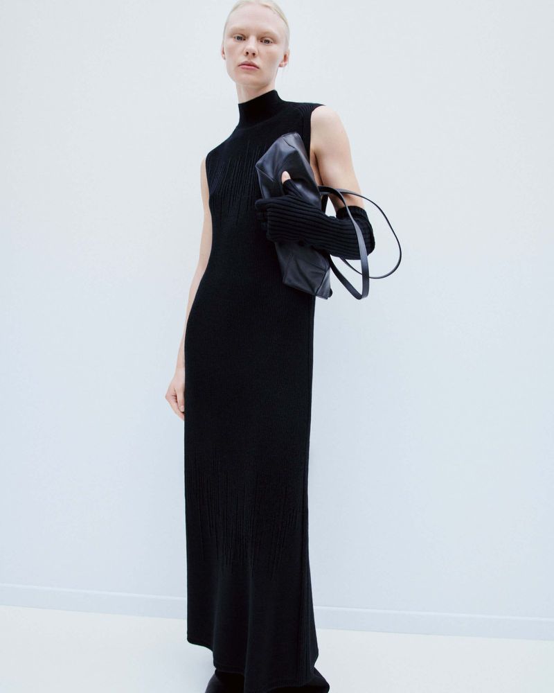 An adult female model poses in a black sleeveless maxi dress, accessorized with gloves and a handbag, against a plain white backdrop.