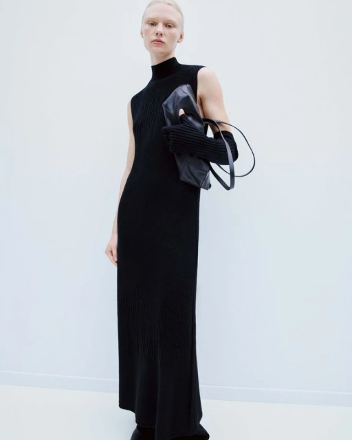 An adult female model poses in a black sleeveless maxi dress, accessorized with gloves and a handbag, against a plain white backdrop.