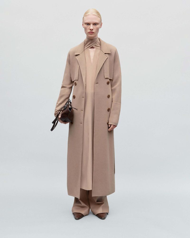 Full-length shot of an adult female model in a tan coat, dress, and trousers, holding a dark brown leather handbag on a neutral background.