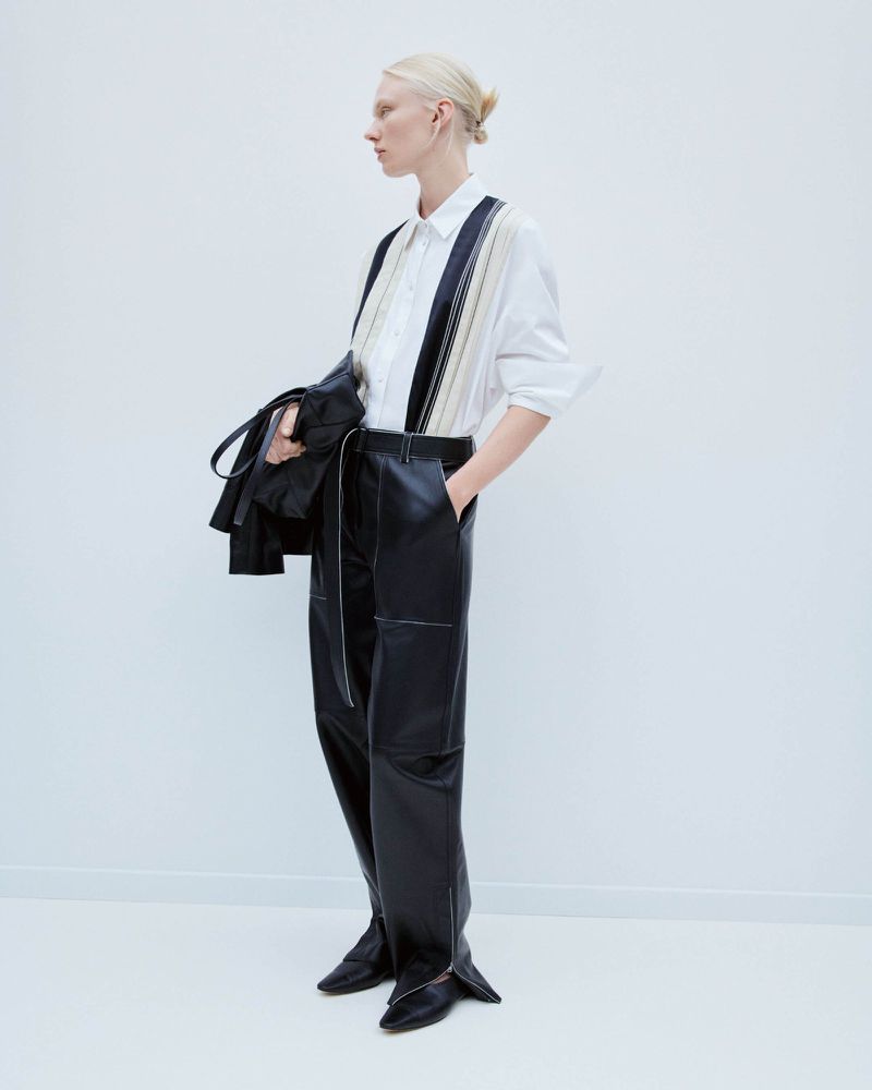 Fashion model showcases a white button-down shirt with striped detailing, paired with high-waisted black leather trousers in a minimalist studio.