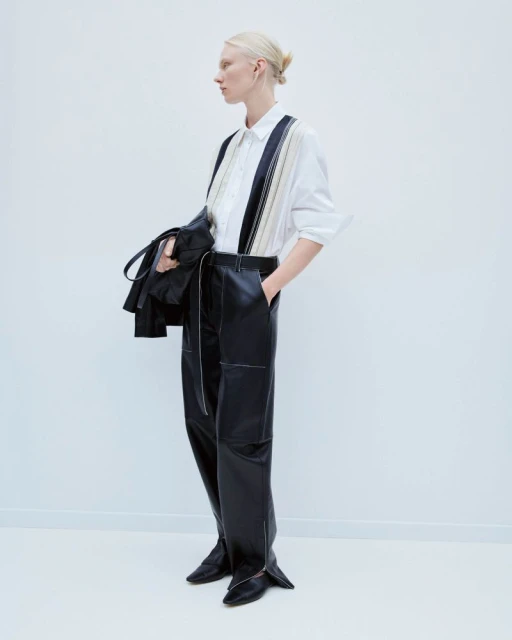 Fashion model showcases a white button-down shirt with striped detailing, paired with high-waisted black leather trousers in a minimalist studio.