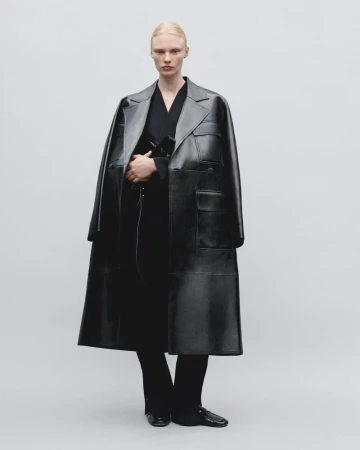 Elegant fashion shot of a woman in a black trench coat, styled with black trousers and loafers, against a neutral backdrop.