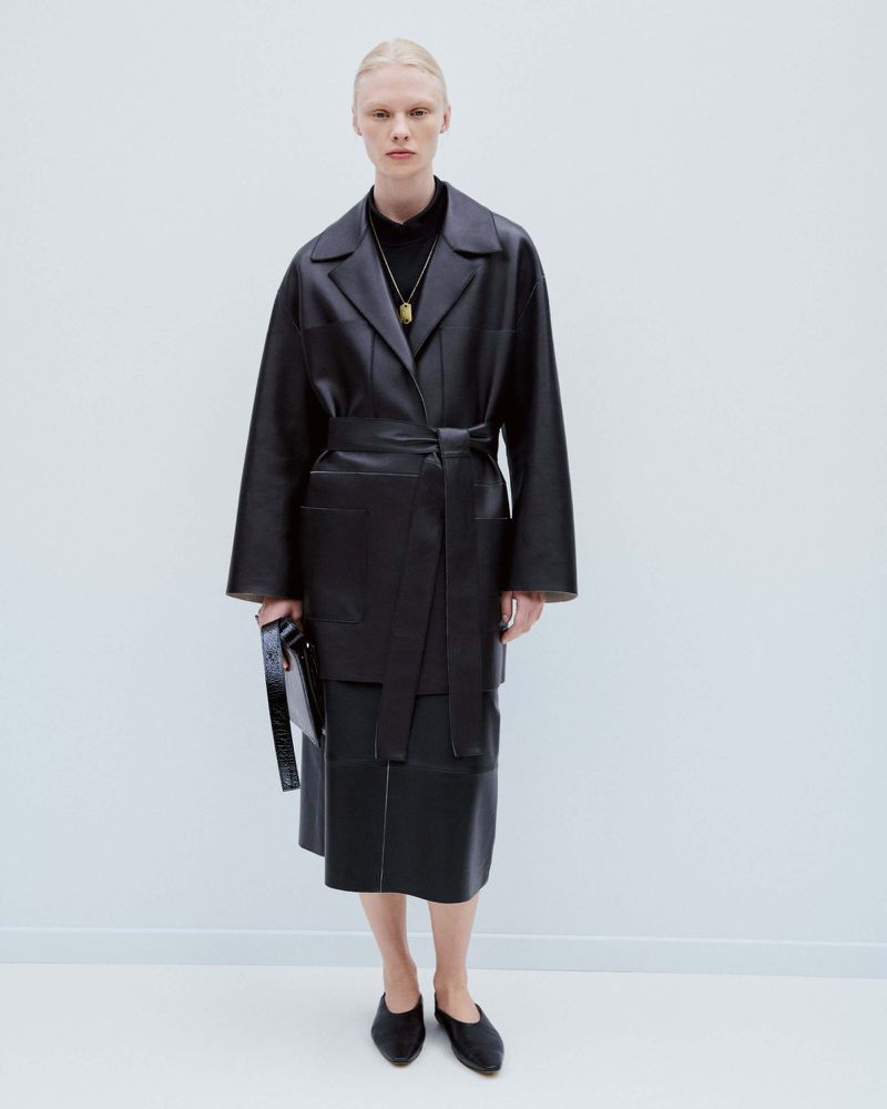 Fashion shot of a woman wearing a black leather belted coat and skirt, standing in a white studio with a neutral expression.