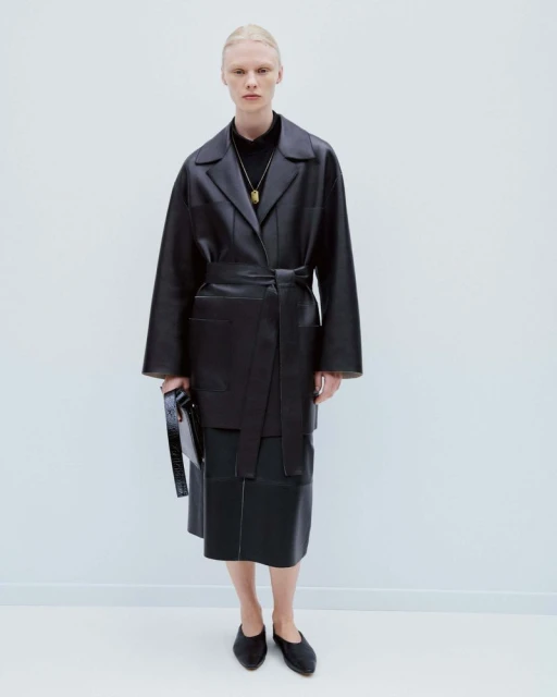 Fashion shot of a woman wearing a black leather belted coat and skirt, standing in a white studio with a neutral expression.