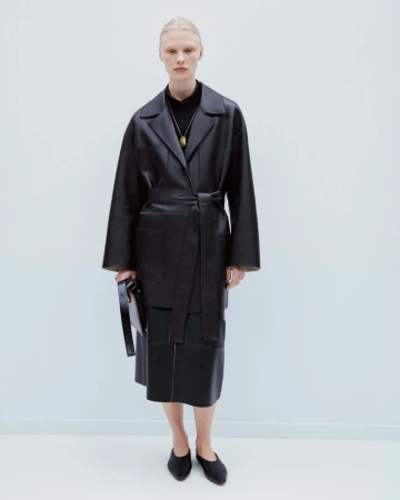 Model in black leather belted coat holding a dark handbag against a seamless white background