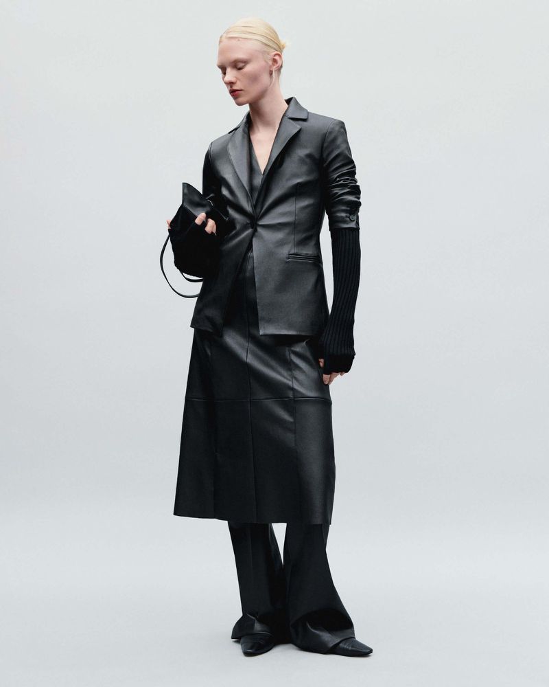 A single female fashion model poses in a black leather blazer, skirt, and fitted long sleeve against a plain, bright backdrop.