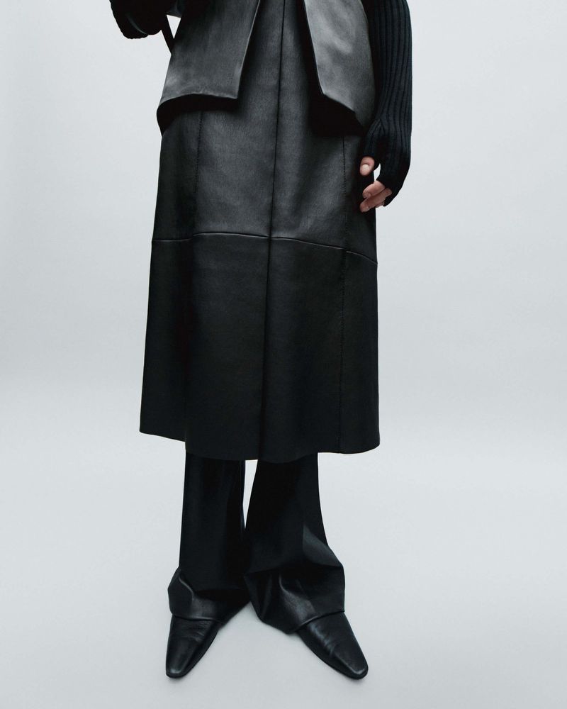 Fashion shot of a woman wearing a long black leather skirt, black sweater and black leather pointy shoes in a studio with gray background.