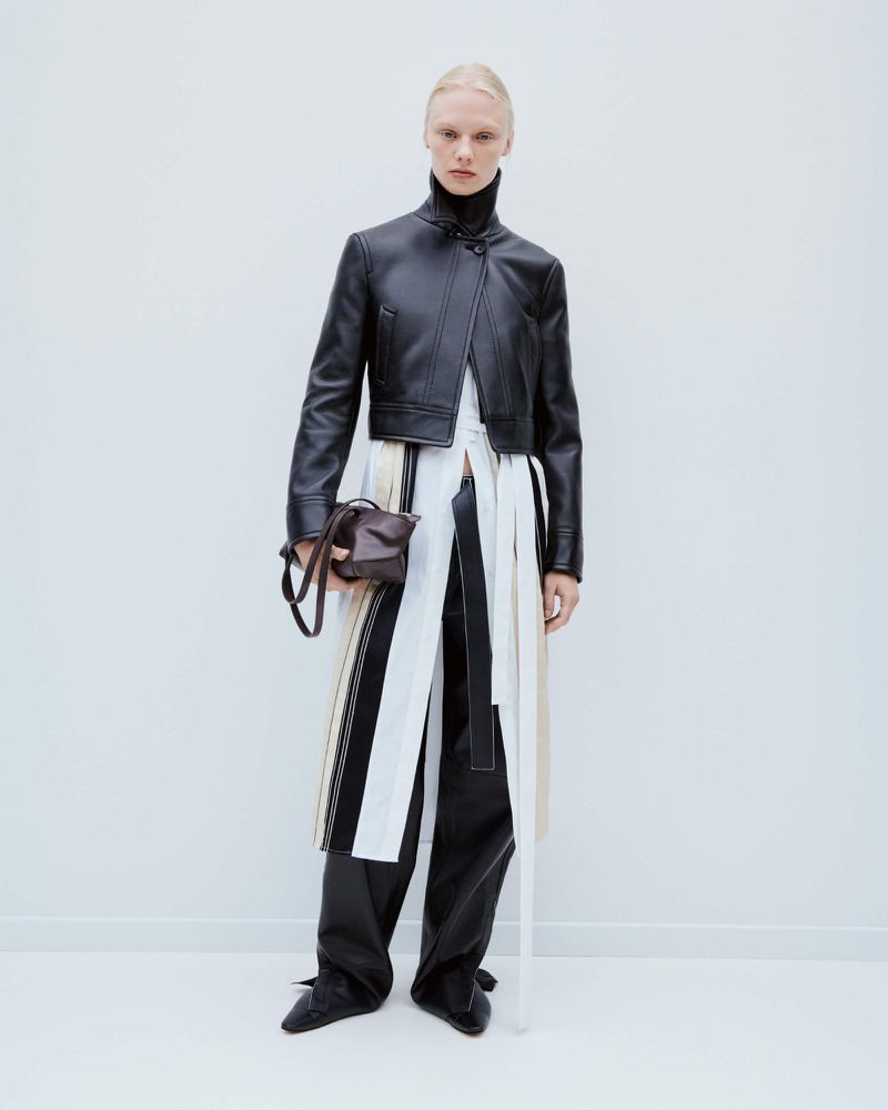 Fashion shot of adult female model in studio wearing a black leather jacket and pants holding a brown handbag, against a white background.
