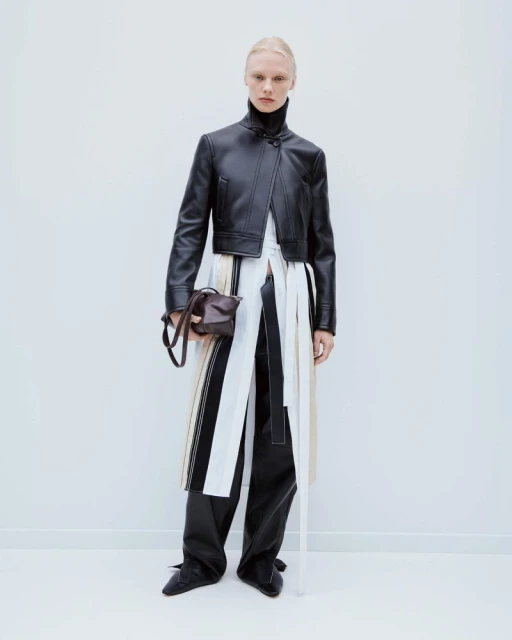 Fashion shot of adult female model in studio wearing a black leather jacket and pants holding a brown handbag, against a white background.