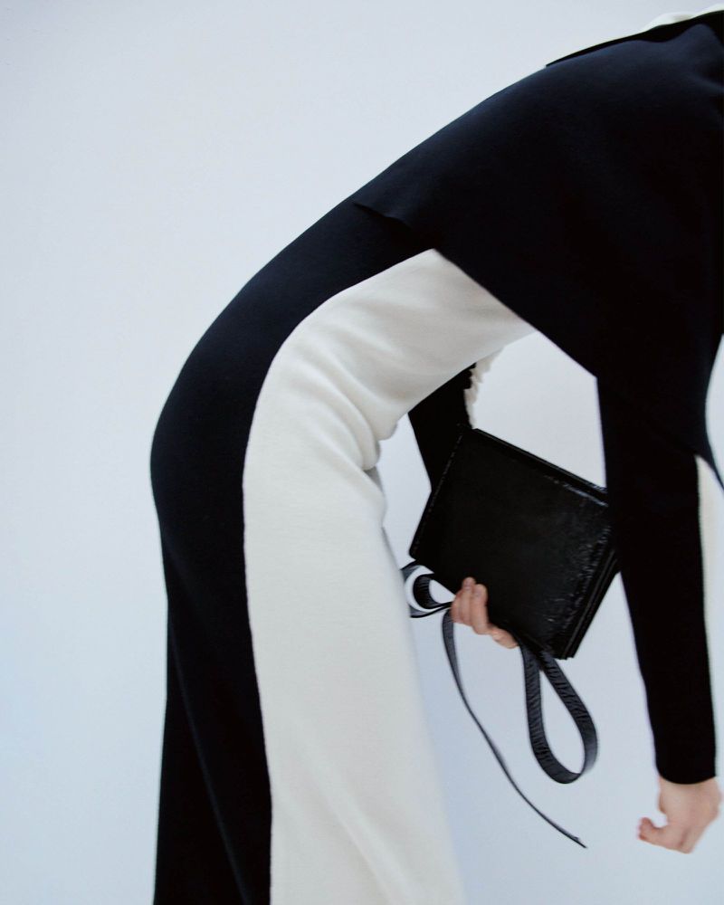 Fashion shoot features a woman in a monochrome jumpsuit holding a black clutch, set against a bright backdrop.