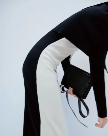 Fashion shoot features a woman in a monochrome jumpsuit holding a black clutch, set against a bright backdrop.