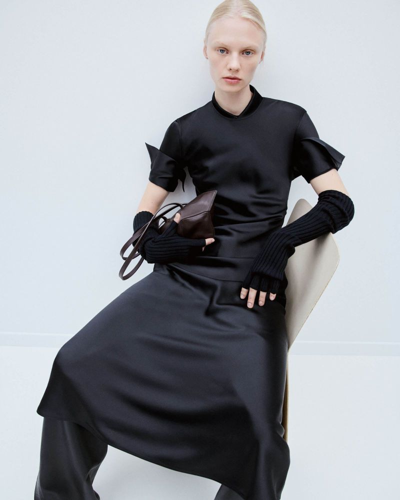 A fair-skinned woman poses seated indoors wearing a black satin dress, black knit arm warmers, and holding a brown leather purse.