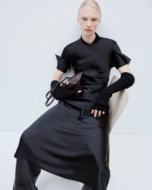 A fair-skinned woman poses seated indoors wearing a black satin dress, black knit arm warmers, and holding a brown leather purse.