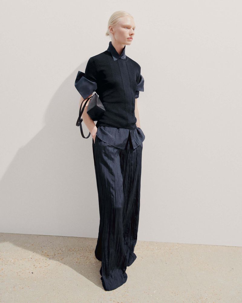 Fashion model in a black top with dramatic sleeves and navy wide-leg pleated pants, accessorized with a small boxy handbag against a white wall.