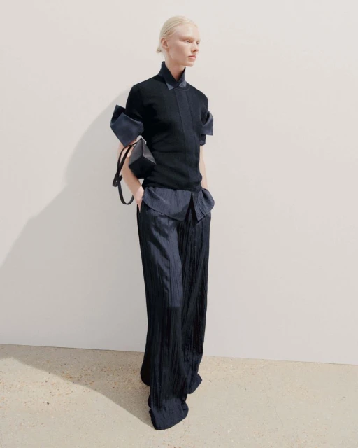 Fashion model in a black top with dramatic sleeves and navy wide-leg pleated pants, accessorized with a small boxy handbag against a white wall.