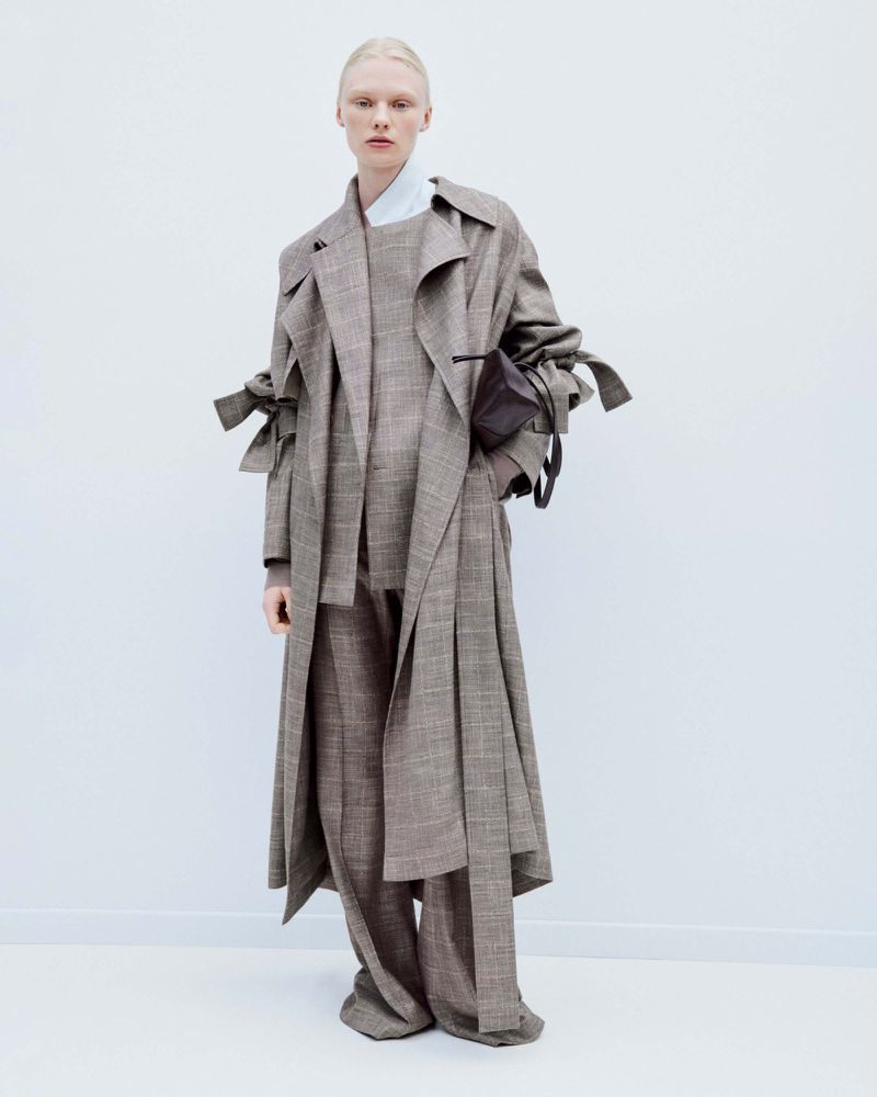 Full-length shot of a fair-skinned person with blonde hair wearing a brown plaid coat, dress and pants in a minimalist indoor studio.