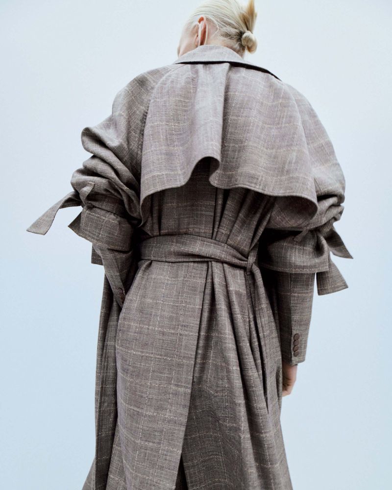 Back view of an adult female with blond hair tied in a bun wearing a brown and beige plaid trench coat with tie belt.