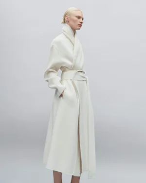 Model wearing a long white belted coat against a neutral grey studio background