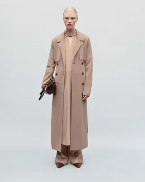 Model standing against a plain light grey backdrop wearing a long beige coat holding a dark handbag