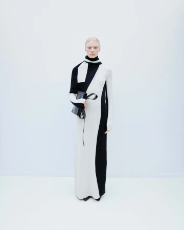 Model wearing a black and white color-blocked turtleneck dress holding a handbag against a plain white seamless background