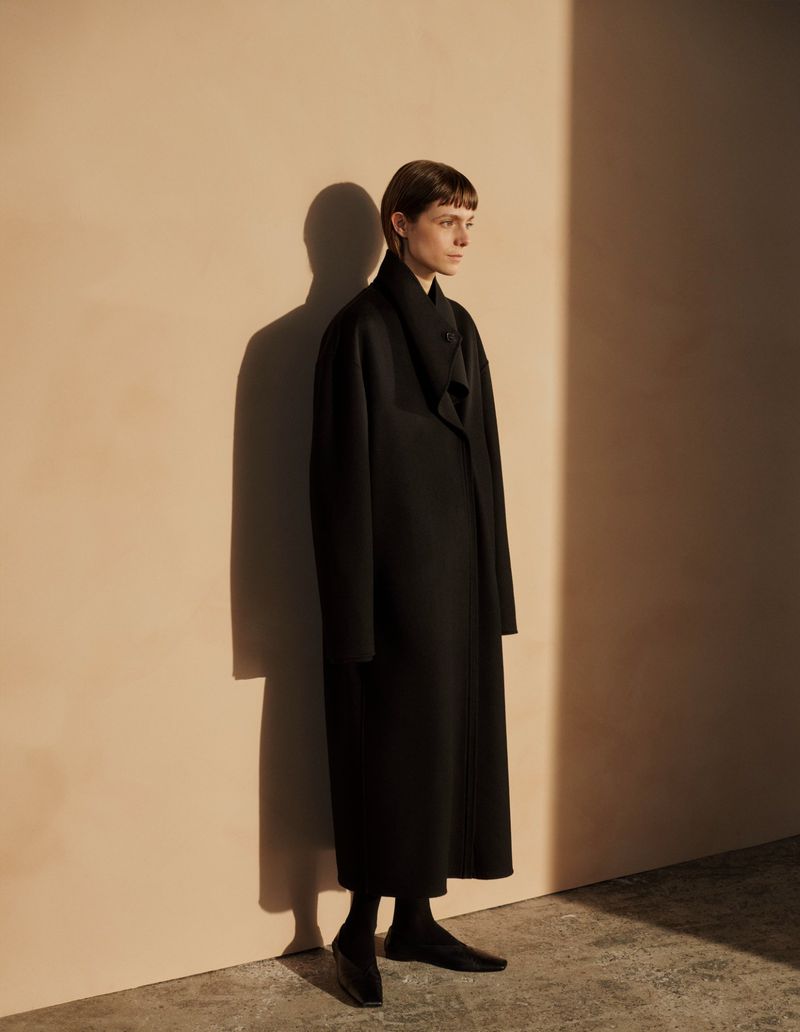 Model wearing a long black coat standing against a warm beige wall with a distinct shadow.