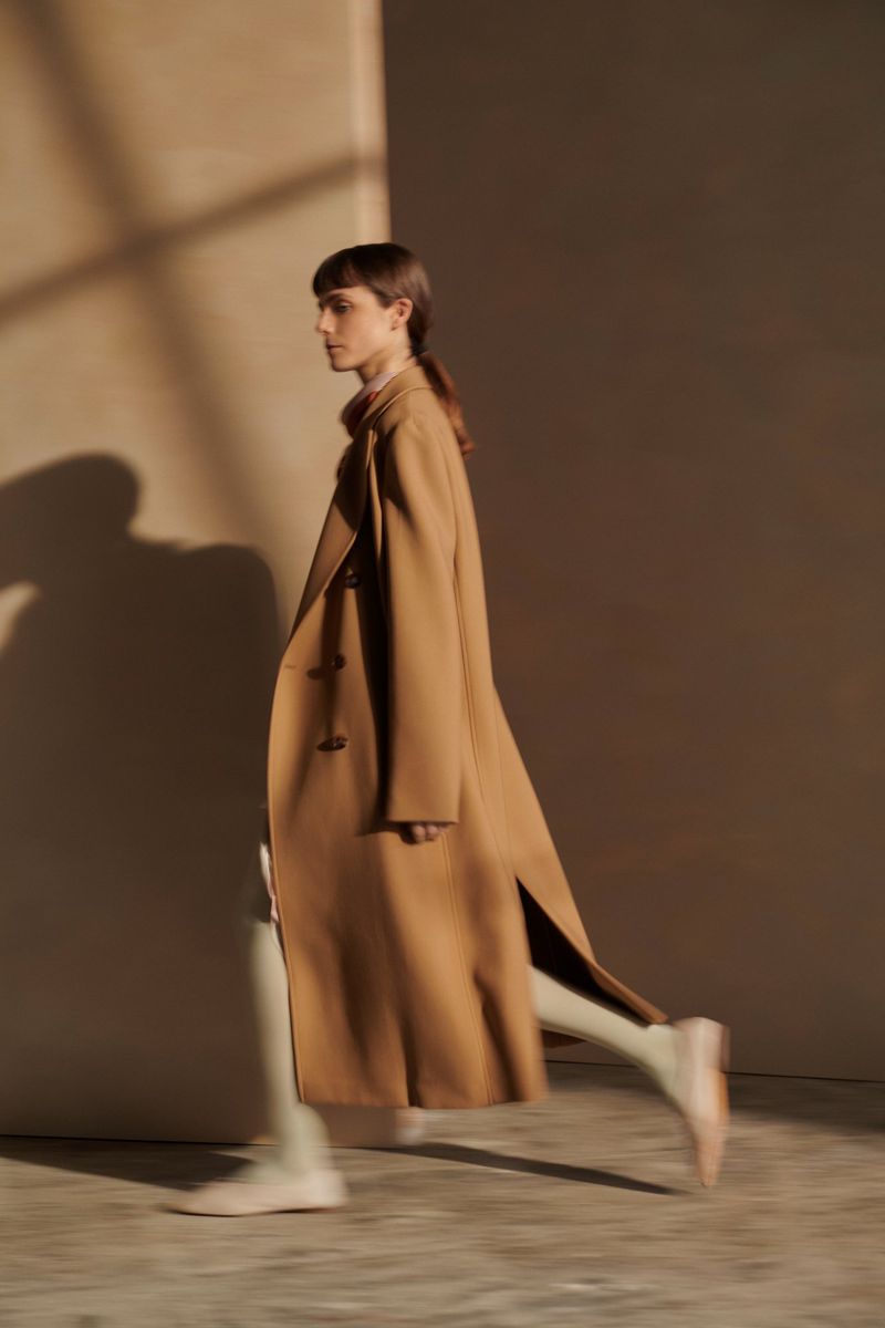 A woman in a tan coat walks against a neutral backdrop, showcasing fashion and movement in a studio or minimalist setting.