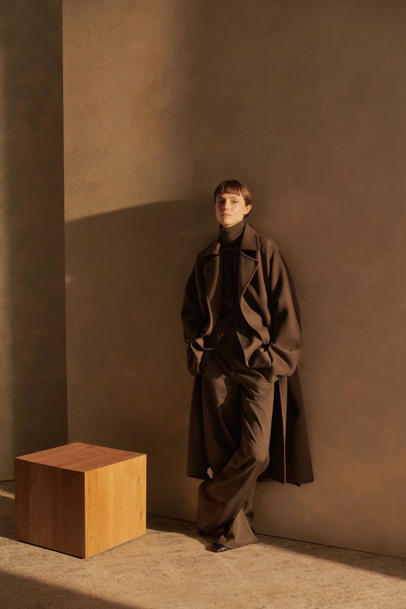 A slender woman with short brown hair wearing a long dark brown coat and matching suit stands against a tan-colored wall.