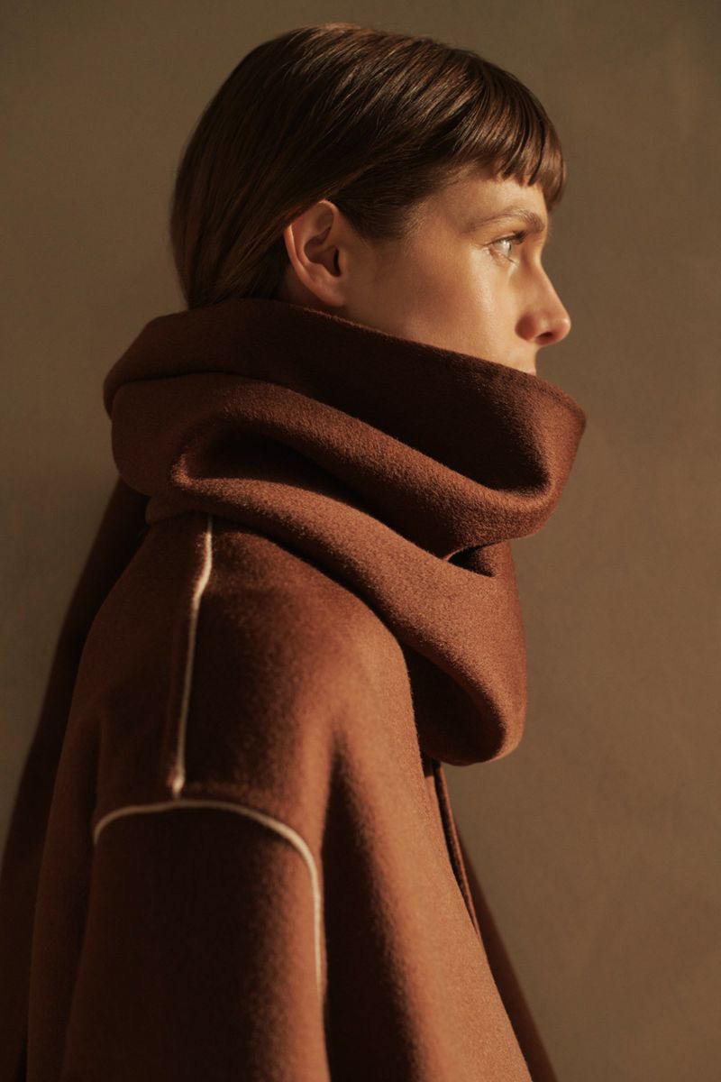 Woman in profile wearing a brown coat and matching scarf wrapped around her neck, set against a neutral background in soft light.