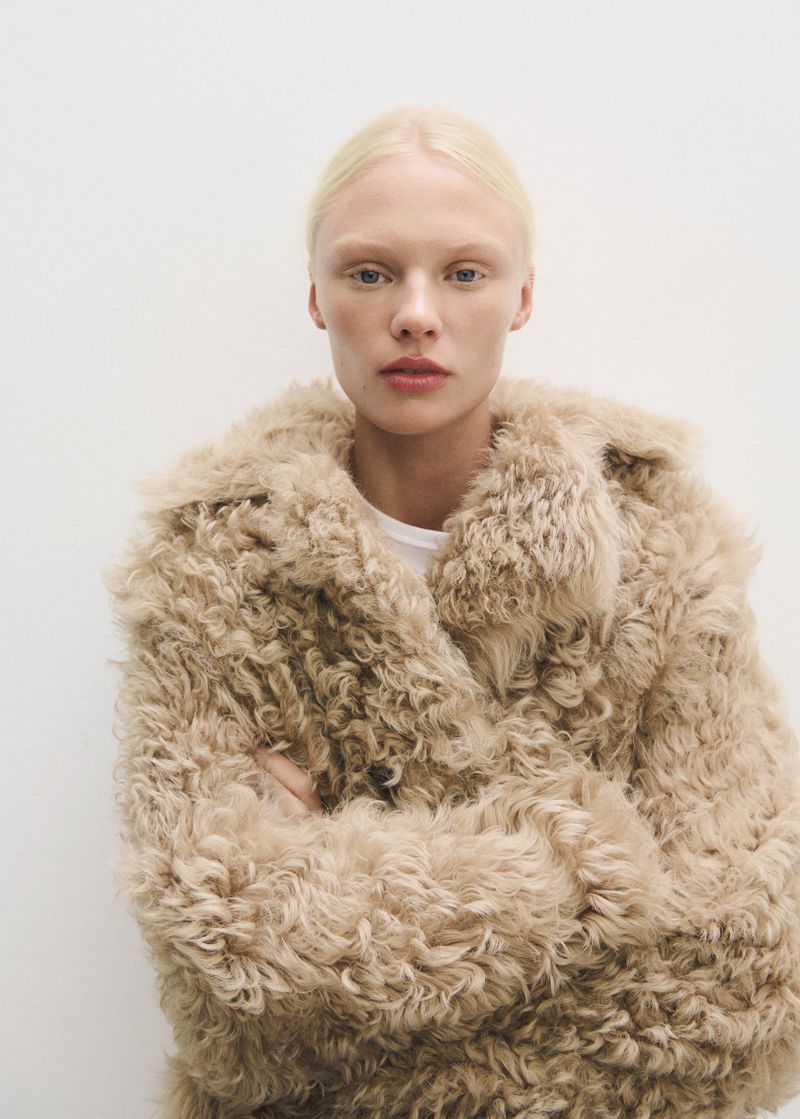 Close-up portrait of a fair-skinned adult woman with blonde hair, wearing a fluffy, light brown coat against a plain white backdrop.