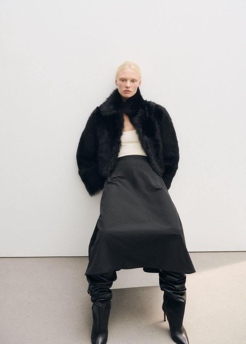 Blonde female model wearing a black fur jacket and dark skirt against a plain studio background.