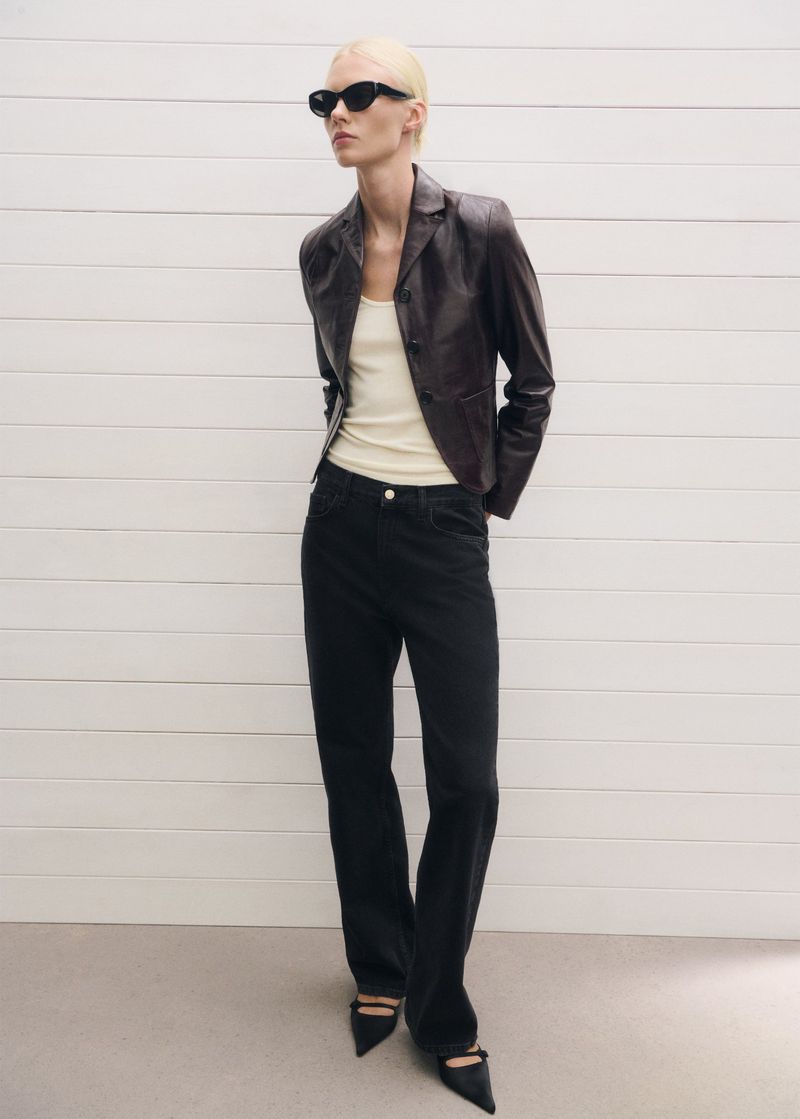 A woman in a brown leather jacket, black jeans, and black sunglasses poses against a white wall in a fashion shot.