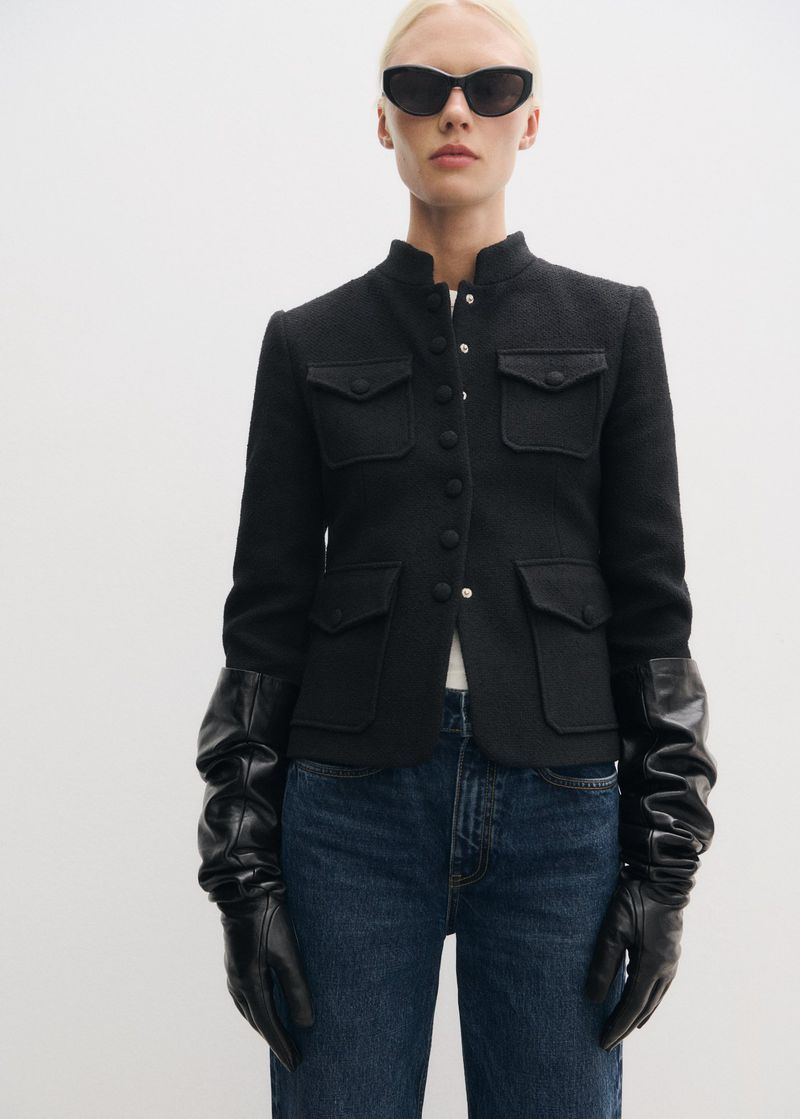 Model wearing black sunglasses, a structured black button-up jacket, and long black leather gloves against white backdrop