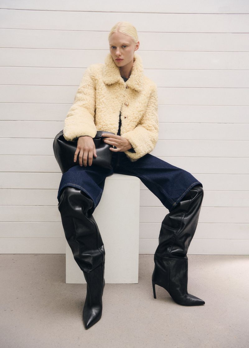 Fashion model wearing teddy jacket, jeans, and thigh-high leather boots sits against a white panel wall in a studio setting.