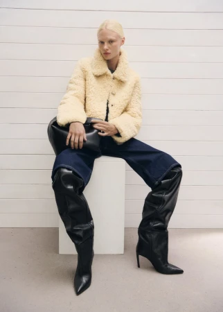 Fashion model wearing teddy jacket, jeans, and thigh-high leather boots sits against a white panel wall in a studio setting.