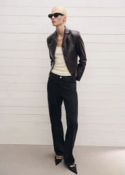Model wearing dark sunglasses, brown leather blazer, cream top, and black jeans against a white paneled wall.