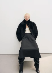 Blonde female model wearing a black fur jacket and dark skirt against a plain studio background.