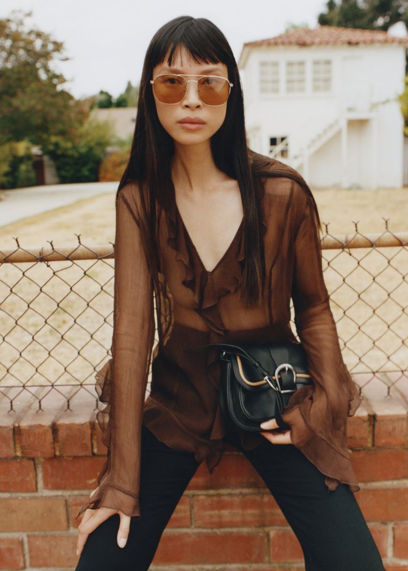 Portrait of a young woman wearing a sheer brown blouse, black pants, sunglasses, and a black purse posing outdoors in front of a wall.