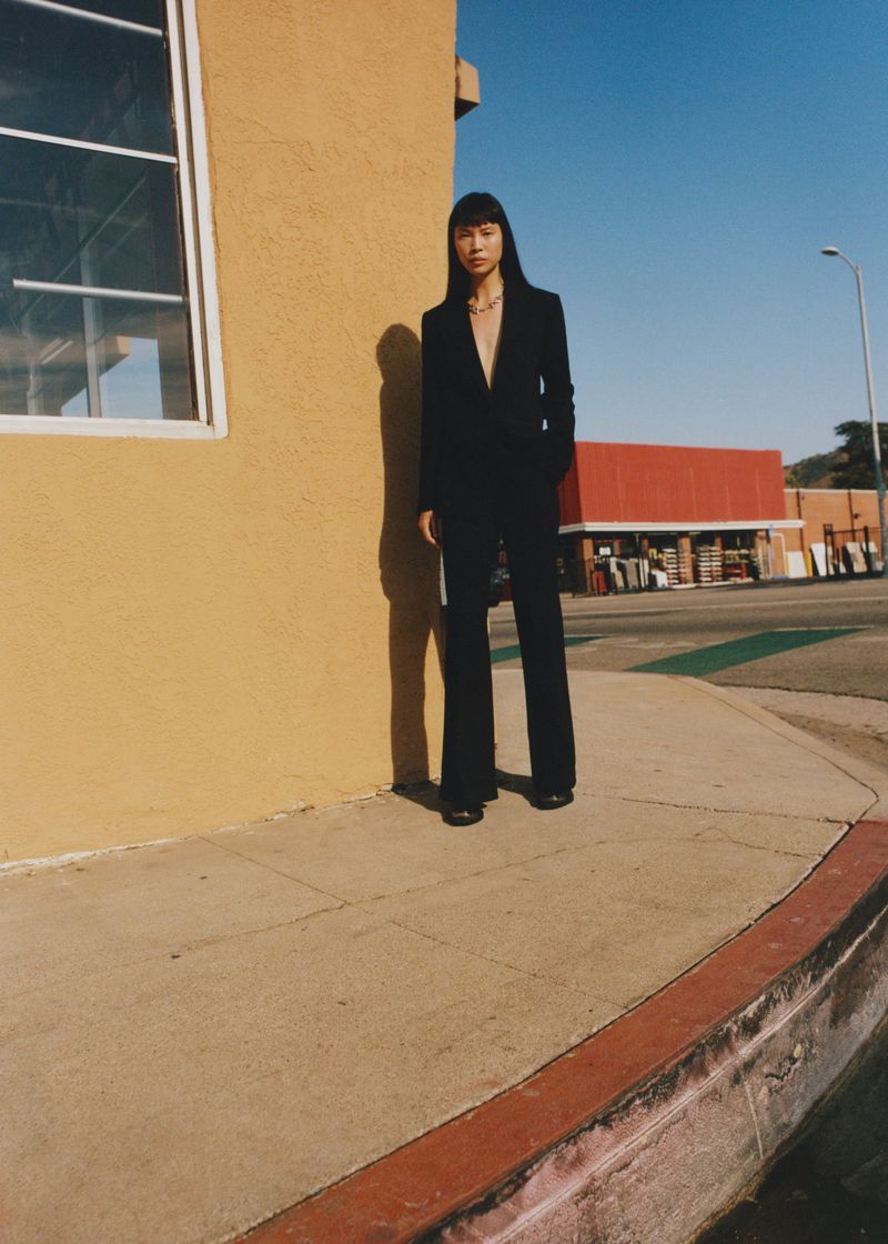 Model standing outdoors wearing a black suit jacket and trousers against a yellow building exterior