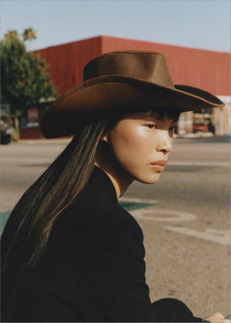 Asian woman in a cowboy hat poses outdoors with a red building and trees in the background on a sunny day, medium shot.