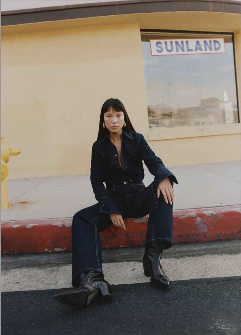 A woman in a dark denim outfit sits on a curb in front of a beige building with a window, wearing black heeled boots in an outdoor fashion shot.