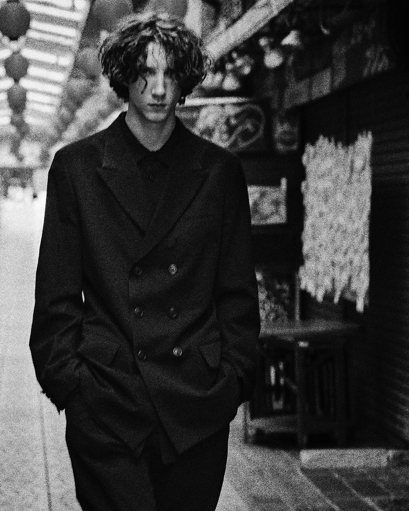 Black-and-white portrait of a young adult male with curly hair wearing a dark double-breasted jacket standing in an urban environment.