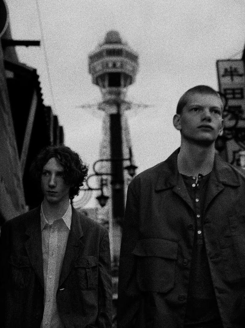 Two young men in jackets stand on a city street in Osaka, Japan, with the Tsutenkaku Tower visible in the background, captured in black and white.