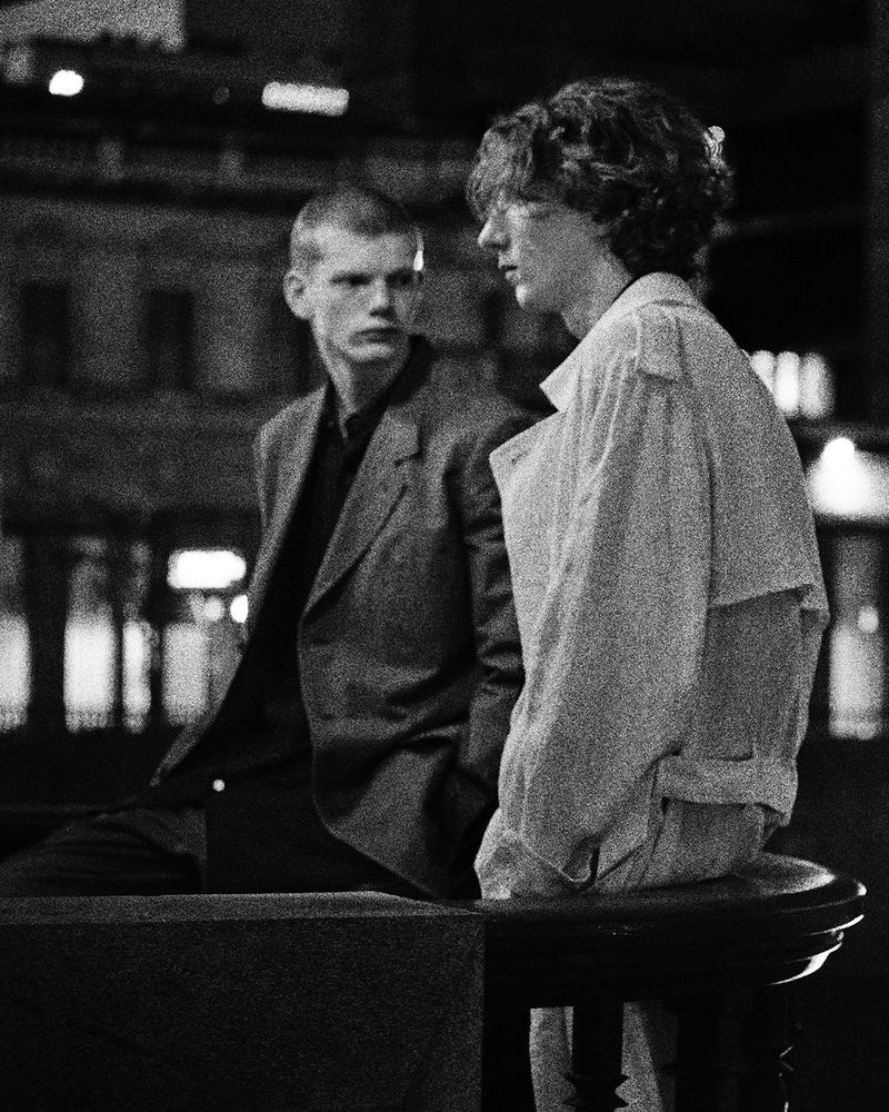 Black and white photo of two men in jackets leaning against a ledge in an urban setting at night, one gazing off to the side.